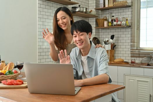 Image of adorable asian couple making video call with friends or family via laptop computer while sitting in cozy kitchen.