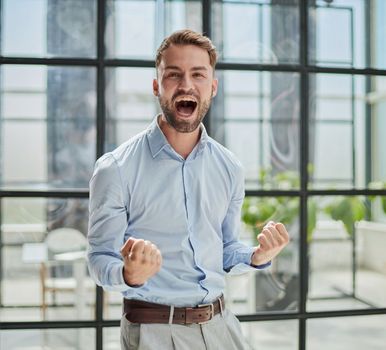 Portrait of a young businessman in a blue shirt shaking his fists celebrating his success in the office