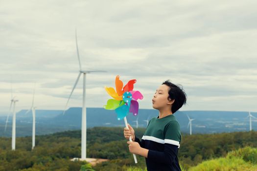 Progressive young asian boy playing with wind pinwheel toy in the wind turbine farm, green field over the hill. Green energy from renewable electric wind generator. Windmill in the countryside concept