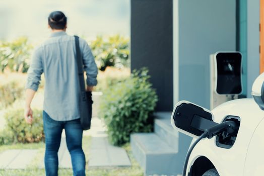 Focus electric car charging at home charging station with blurred progressive man walking in the background. Electric car using renewable clean for eco-friendly concept.