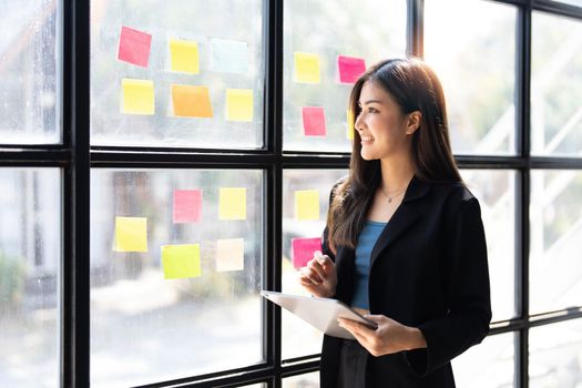 Young creative woman professional using sticky notes in glass wall to writing strategy business plan to development grow to success. Happy asian woman working at office...