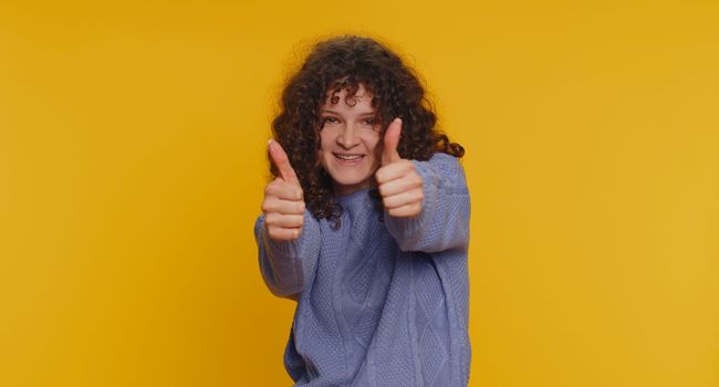 Like. Pretty curly haired woman in sweater raises thumbs up agrees with something or gives positive reply recommends advertisement likes good. Young girl isolated alone on yellow studio background