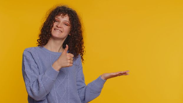 Lovely woman showing thumbs up and pointing empty place, advertising area for commercial text, copy space for goods promotion. Young curly haired girl. Indoor studio shot on yellow wall background