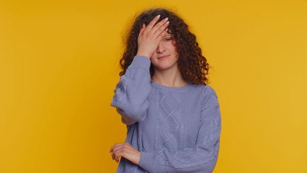 Face palm. Shame on you. Upset curly haired woman making face palm gesture, feeling bored, disappointed in result, bad news, ridiculous joke. Young teen girl isolated alone on yellow studio background