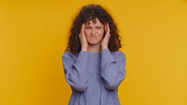 Hipster young curly haired woman in sweater rubbing temples to cure headache problem, suffering from tension, migaine, stress, grimacing in pain, high blood pressure isolated on yellow wall background