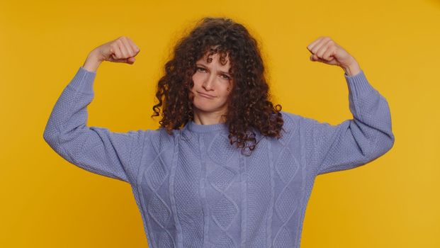 I am strong and independent. Curly haired woman showing biceps, looking confident, feeling power strength to fight for rights, energy to gain success win. Young teen girl on yellow studio background
