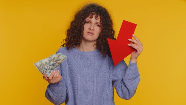 Upset curly haired woman showing red arrow pointing down, concept of downgrade, unsuccessful business, fall of stock market money dollar exchange rate bankruptcy fail. Young girl on yellow background