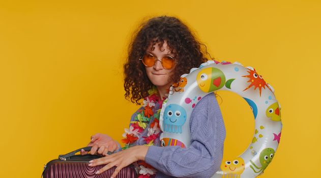 Happy curly haired woman in sunglasses, hawaiian lei walking along with luggage bag, swimming ring. Young smiling girl dancing, on yellow studio background. Summer vacation, journey, trip to seaside