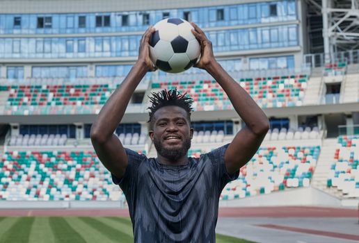 Young confident african american guy holds soccer ball in hands outside, on stadium and looks up.