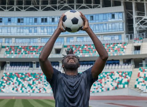 Young confident african american guy holds soccer ball in hands outside, on stadium and looks up.