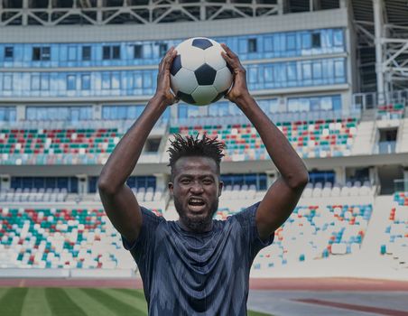 Young confident african american guy holds soccer ball in hands outside, on stadium and looks up.