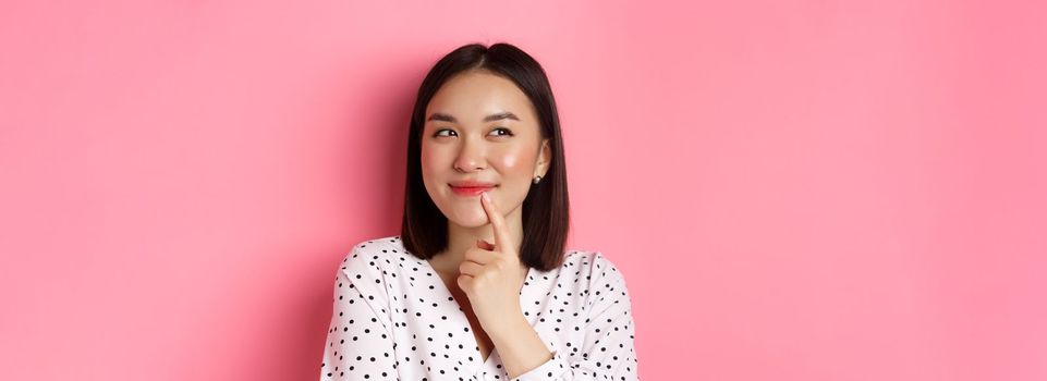 Beauty and lifestyle concept. Close-up of dreamy asian girl smiling, looking left and thinking, making choice, standing over pink background.