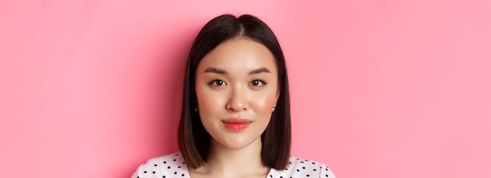 Beauty and skin care concept. Headshot of young feminine asian woman with short hairstyle, looking at camera, standing over pink background.