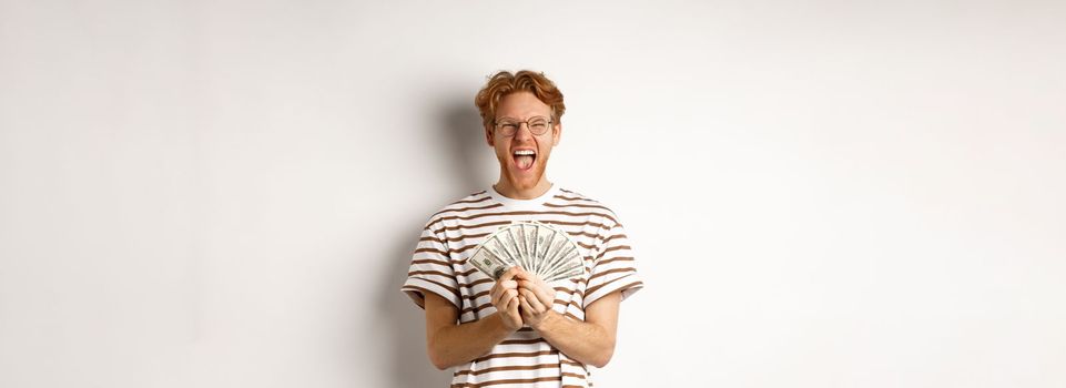 Lucky young man with red hair showing dollars, winning money and screaming of happiness, holding prize cash, standing over white background.