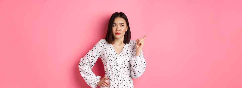 Beautiful asian girl pointing finger at upper left corner, looking confused and doubtful, standing over pink background.
