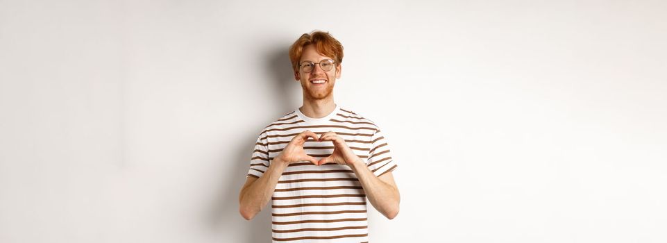 Valentines day. Happy boyfriend with red hair, smiling and showing heart gesture, I love you, standing over white background.