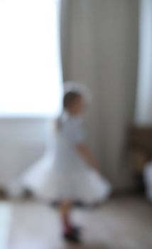 little dancing ballerina girl out of focus indoors. Little Caucasian girl in babydoll dress standing by big window. Image with selective focus.