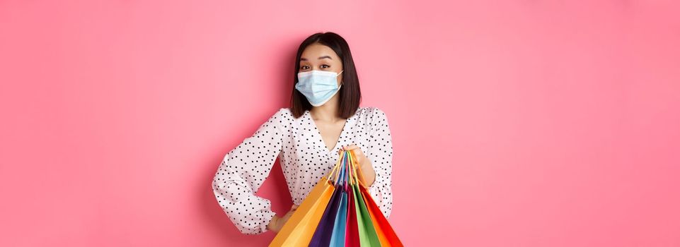 Covid-19, pandemic and lifestyle concept. Beautiful asian woman in medical mask going shopping, holding bags and smiling with eyes, standing over pink background.