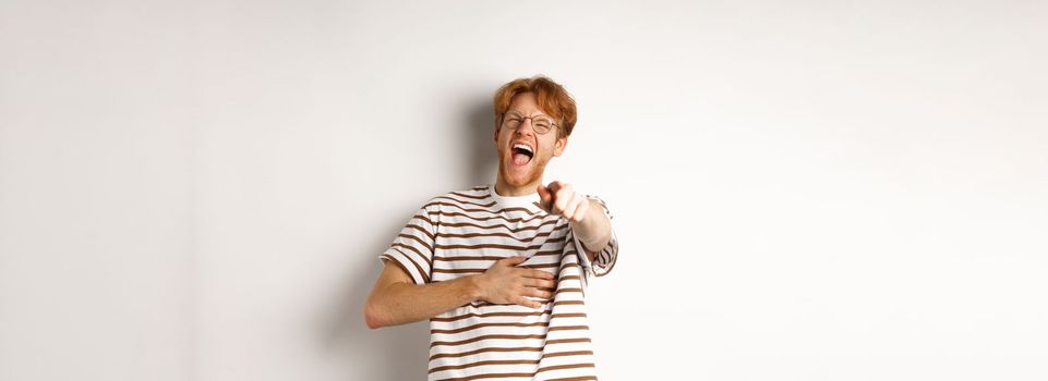 Young man with ginger hair and beard pointing finger at camera and laughing, making fun of someone hilarious, standing over white background.