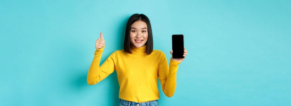 Technology and people concept. Cheerful asian girl in yellow sweater showing blank smartphone screen and thumbs up, demonstrate online offer, standing over blue background.