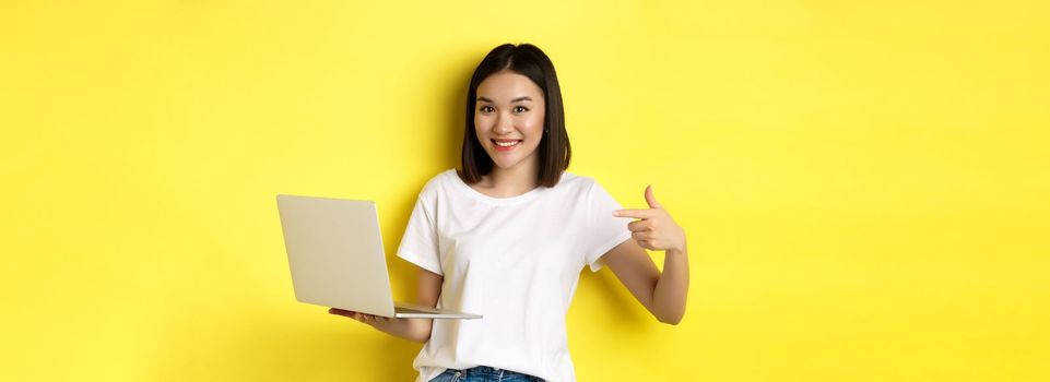 Smiling asian woman pointing finger at her laptop, showing something online, standing over yellow background.