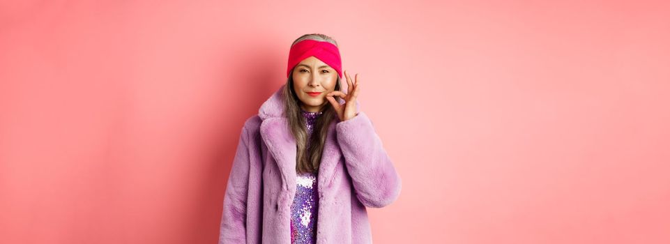 Fashion and shopping concept. Cunning asian senior woman in stylish clothes keeping secret, squinting and looking devious at camera, zipping mouth, making promise, pink background.