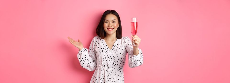 Beautiful asian woman raising glass of champagne and looking happy at camera, congratulating you on party, celebrating, standing over pink background.