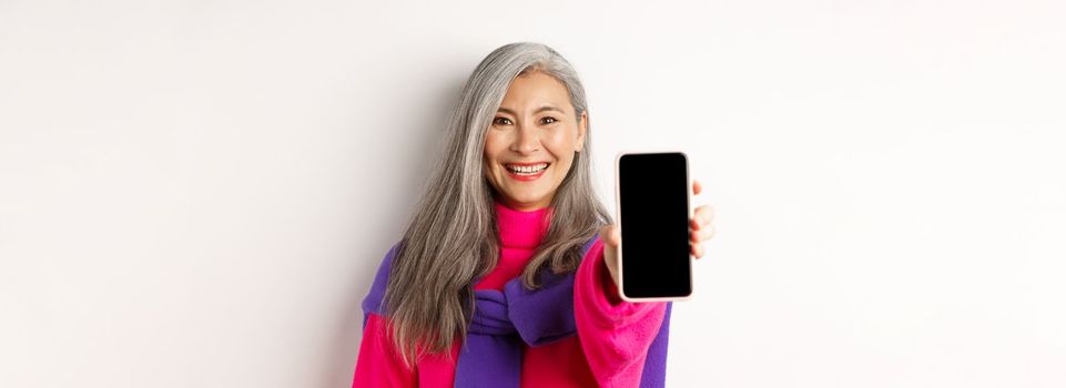 Online shopping. Close up of modern asian senior woman extending hand with mobile phone, showing blank smartphone screen and smiling, standing over white background.