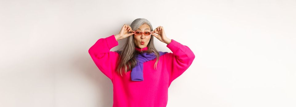 Trendy asian senior woman in sunglasses, pucker lips and looking surprised at camera, staring amazed at special offer, standing over white background.