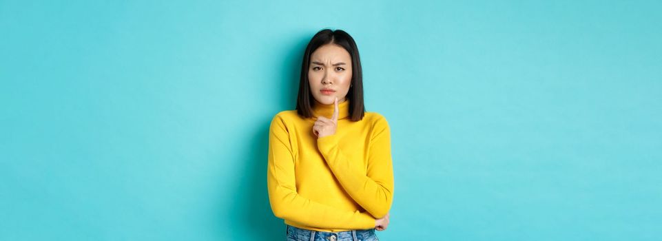 Image of pensive asian woman touching lip and frowning, thinking about something, trying to understand, standing over blue background.