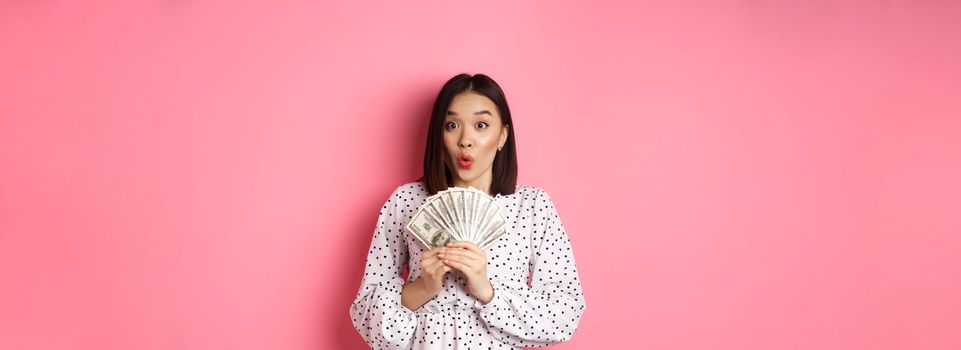Shopping concept. Excited asian woman holding money, gasping amazed and staring at camera, standing over pink background.
