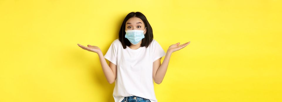 Covid-19, quarantine and social distancing concept. Clueless asian woman in medical mask shrugging shoulders, spread hands sideways clueless, know nothing, yellow background.