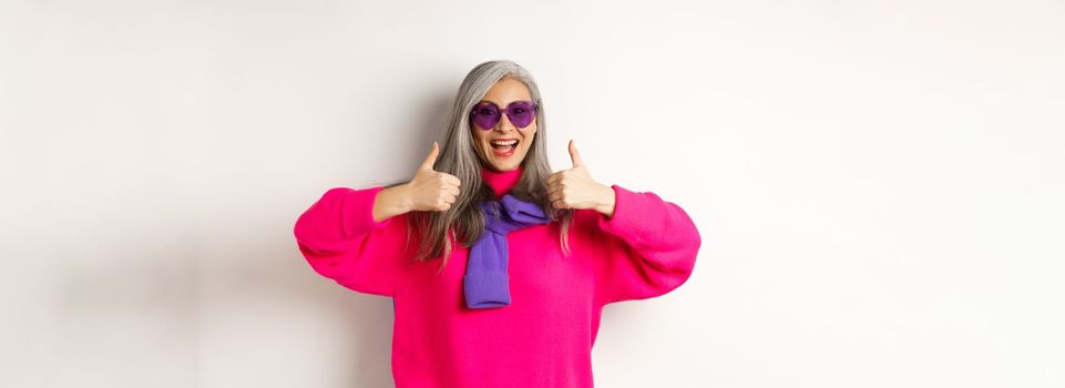 Shopping concept. Stylish asian senior woman in sunglasses and trendy outfit, showing thumb-up in approval, recommending shop, standing over white background.