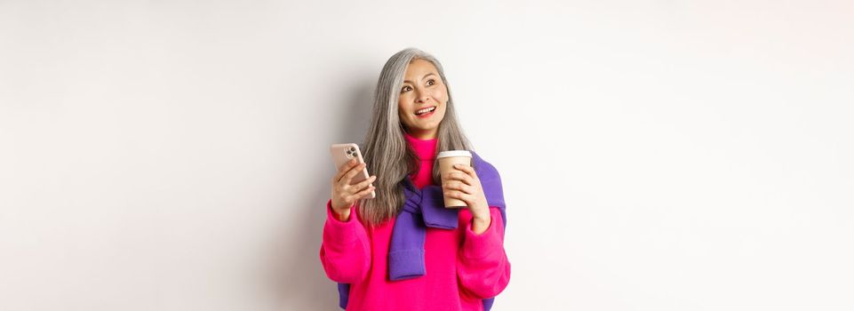 Fashionable asian mature woman with grey hair, drinking coffee and using smartphone, looking upper left corner promotion with dreamy face, white background.