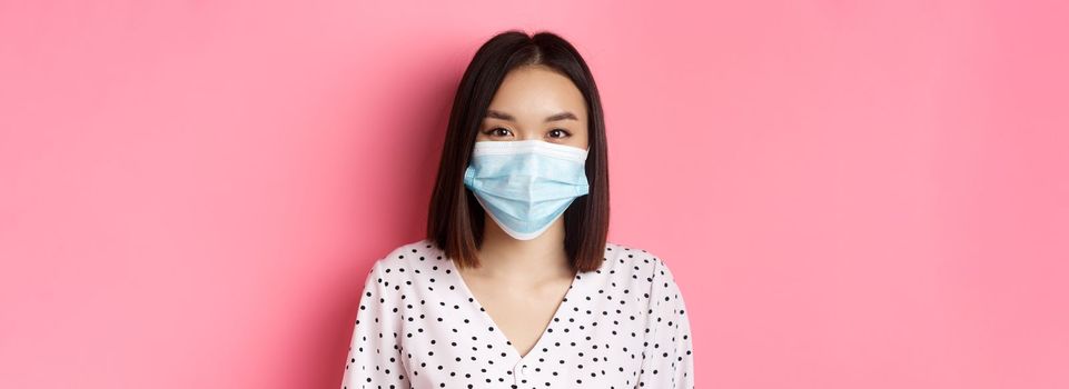 Covid-19, pandemic and lifestyle concept. Close-up of beautiful asian woman in face mask smiling with eyes, wearing protection from coronavirus in public, pink background.