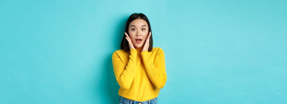 Portrait of surprised asian girl checking out promo, gasping amazed and touching cheeks, staring at camera amazed, blue background.