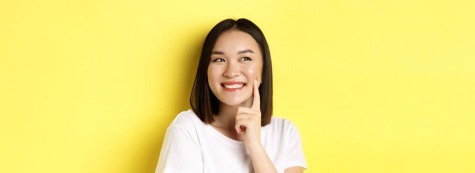 Beauty and skincare. Close up of young asian woman with short dark hair, healthy glowing skin, smiling and touching dimples on cheeks, standing over yellow background.
