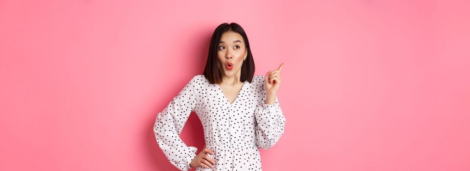 Amazed asian girl in dress pointing finger at upper left corner promo offer, looking interested, shopping and standing over pink background.
