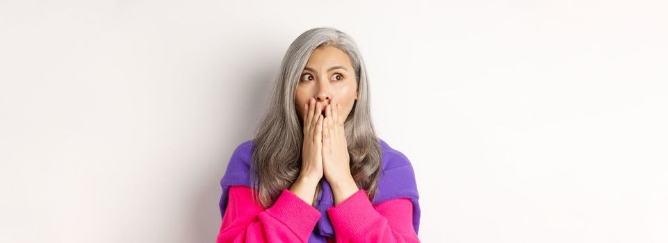 Close-up of shocked asian middle-aged woman with grey hair, gasping and covering mouth startled, looking left, realising something, white background.