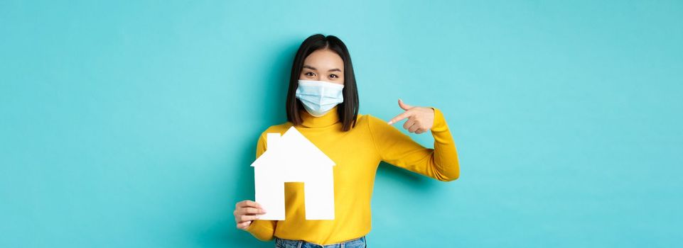 Covid-19, pandemic and real estate concept. Cheerful asian woman smiling in medical mask, showing paper house cutout, recommend agency for buying property, blue background.