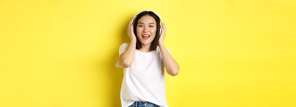 Attractive asian woman enjoying listening music in wireless headphones, smiling pleased and singing along, yellow background.