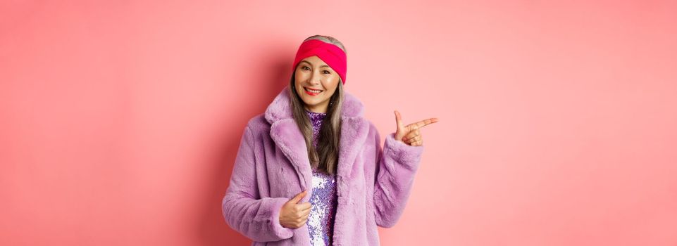 Fashion and shopping concept. Stylish senior asian woman pointing finger left at promotion deal, smiling at camera, suggesting special offer, standing against pink background.