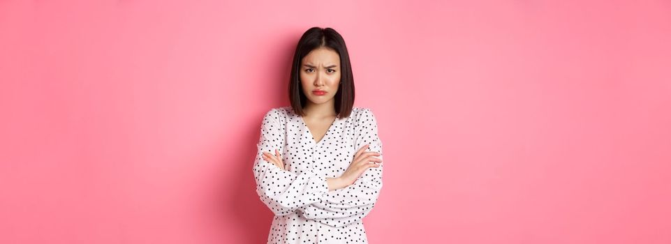 Timid and offended asian girl cross arms on chest, staring defensive and insulted at camera, standing in dress over pink background.