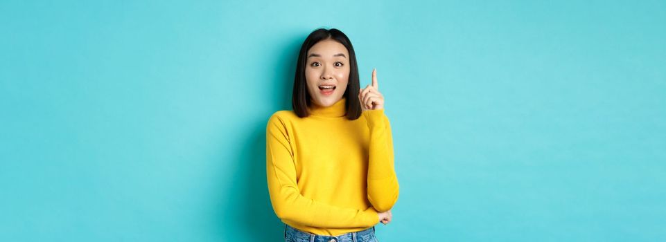 Excited asian woman with short dark hair, pitching an idea, raising finger in eureka gesture and smiling, standing over blue background.