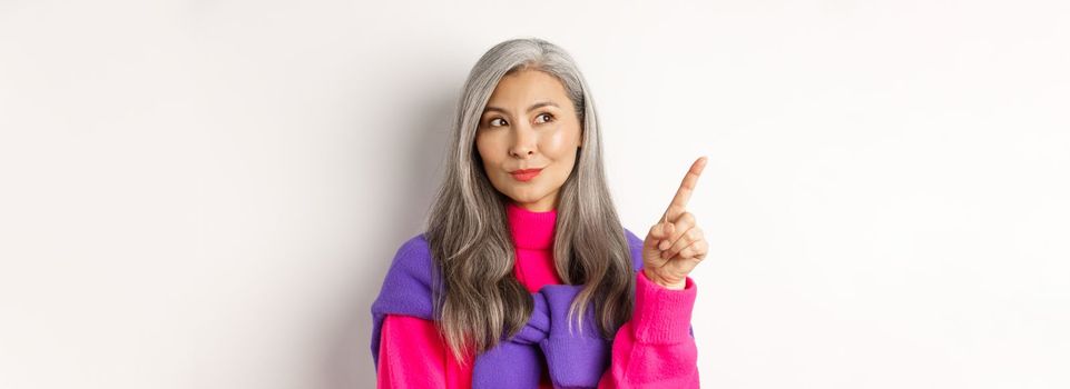 Close-up of stylish korean senior woman with grey hair looking and pointing upper left corner, showing promotion offer, standing over white background.