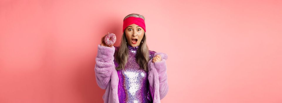 Fashion and food concept. Beautiful asian senior woman in stylish glittering dress holding sweet donut and screaming at camera, scared of calories, pink background.