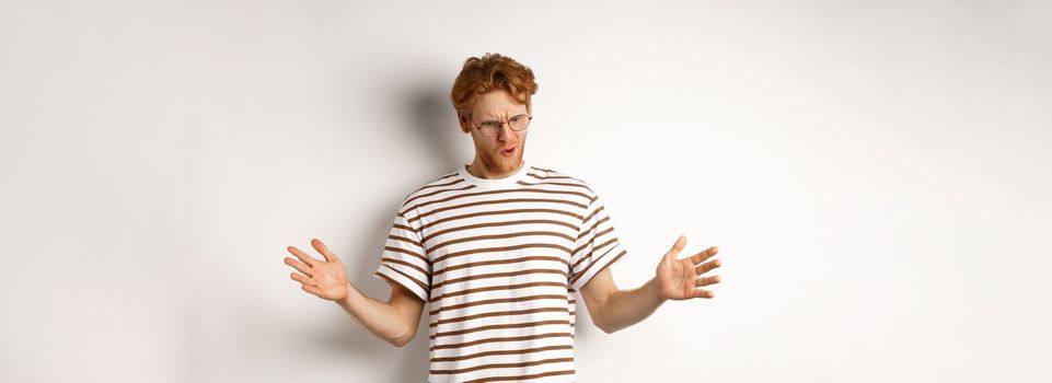Impressed redhead man in glasses showing length of something big, demonstrate large size and looking down, standing over white background.