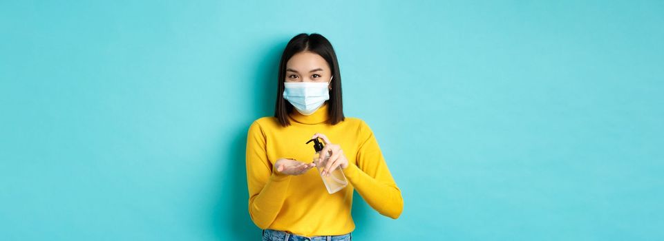 Covid-19, social distancing and pandemic concept. Smiling asian woman in medical mask using preventive measures from coronavirus, using hand sanitizer, standing over blue background.