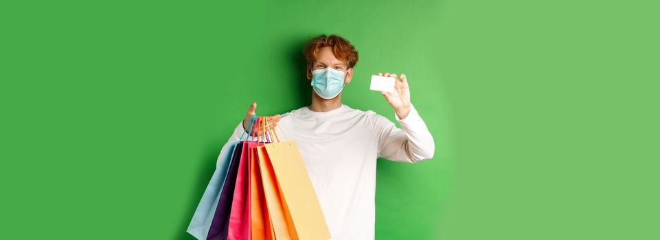 Handsome young man in medical mask, showing plastic credit card and shopping bags with items purchased with discount, green background.