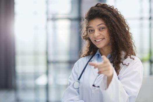 Smiling young female doctor in white coat standing and pointing forward with finger